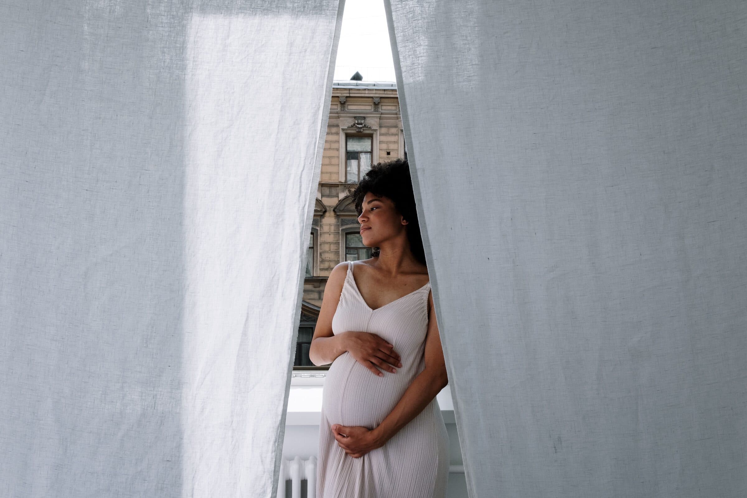 A pregnant woman standing between white curtains at an open window
