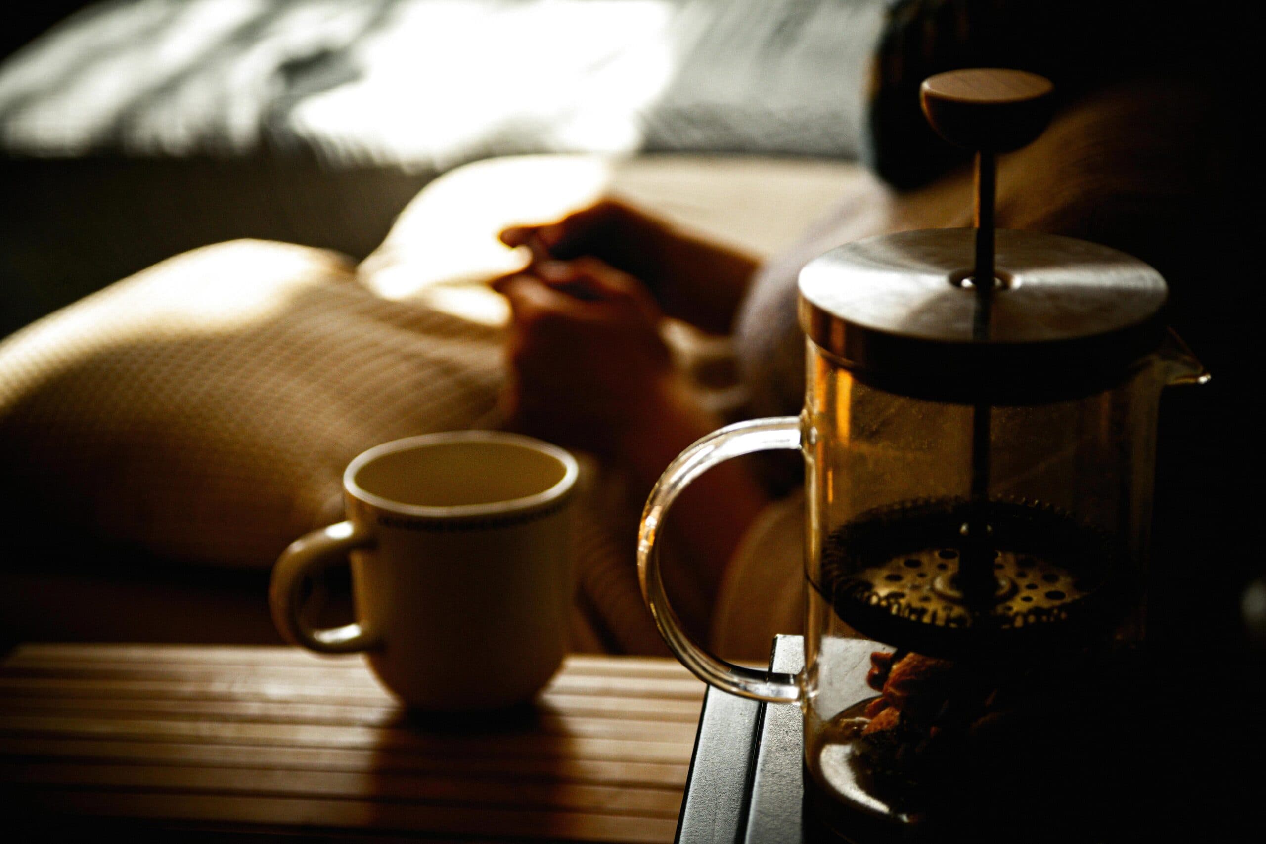 A mug and a cafetiere on a tray, with a woman resting in bed behind them in low light