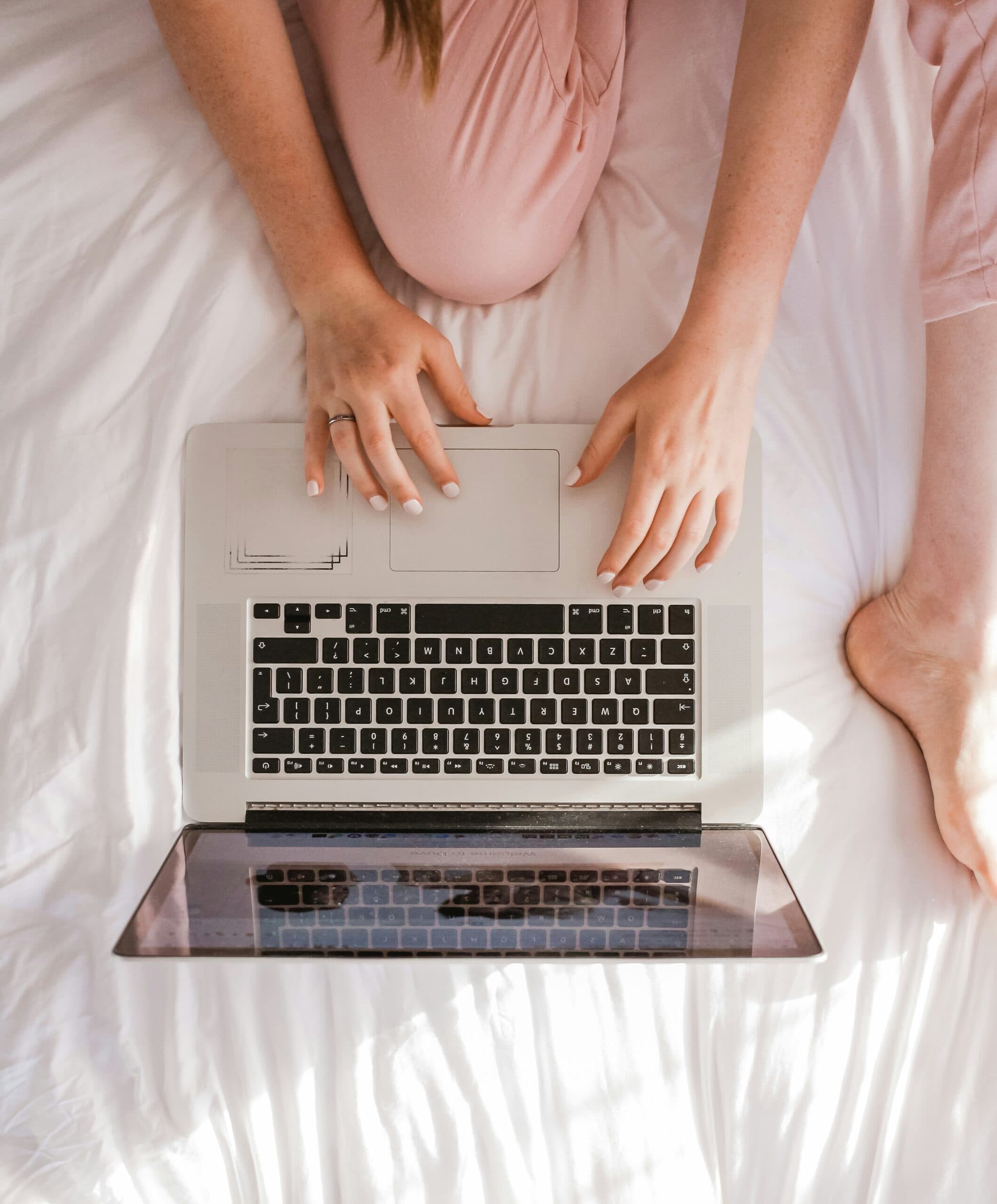 A pregnant woman in soft pink, sitting on a bed with a laptop, ready for a virtual session
